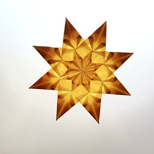 Könnte beinhalten: Ein goldener, achtspitziger Stern aus gefaltetem Papier. Der Stern hat ein geometrisches Design mit mehreren dreieckigen Facetten, wodurch ein strukturiertes Aussehen entsteht. Der Stern ist vor einem weißen Hintergrund zentriert.