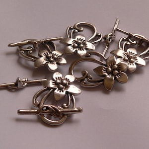 Könnte beinhalten: Silberfarbene Metallschließen mit floralen Designs. Jede Schließe zeigt eine zarte Blume mit einem zentralen Detail und filigranen Ranken.