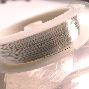 Può includere: Un rocchetto di filo color argento avvolto attorno a una bobina di plastica bianca.