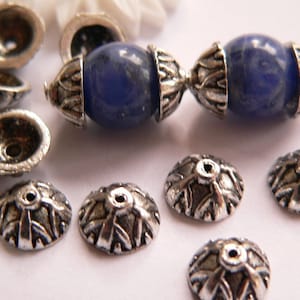 Può includere: Un set di cappucci per perline in metallo argentato con un design decorativo, insieme a due perline ovali con un motivo a marmo blu e bianco.