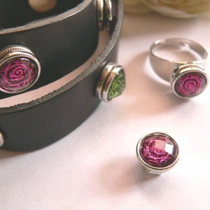 Könnte beinhalten: Ein schwarzes Lederarmband mit silbernen Akzenten und drei rosa und grünen floralen Verzierungen. Das Armband ist zweimal um sich selbst gewickelt. Es gibt auch einen silbernen Ring mit einer rosa floralen Verzierung.