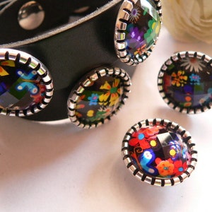 Könnte beinhalten: Ein schwarzes Lederarmband mit vier bunten, runden Glascabochons, die in silberne Zargen gefasst sind. Die Cabochons zeigen florale und geometrische Muster.