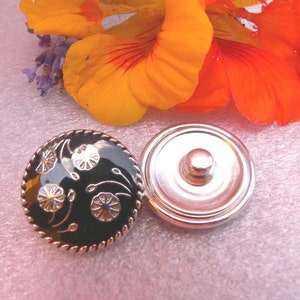 Könnte beinhalten: Ein schwarzer und silberner Druckknopf mit floralem Design. Der Knopf hat einen schwarzen Hintergrund mit silbernen Blumen und Stielen.