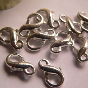 Può includere: Una collezione di chiusure a moschettone argentate. Queste chiusure sono curve con una leva a molla per una chiusura sicura. Sono comunemente utilizzate nella creazione di gioielli per collane, bracciali e altri lavori artigianali. Le chiusure sono mostrate su uno sfondo chiaro.