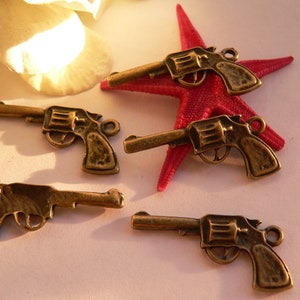 Könnte beinhalten: Fünf antike Revolver-Anhänger in Goldton. Die Anhänger sind detailliert und haben ein realistisches Aussehen. Die Anhänger werden auf einem roten Seestern-Hintergrund präsentiert.