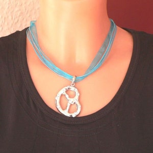 Peut inclure: Un collier en ruban turquoise avec un pendentif en forme de bretzel argent&eacute; incrust&eacute; de strass transparents.