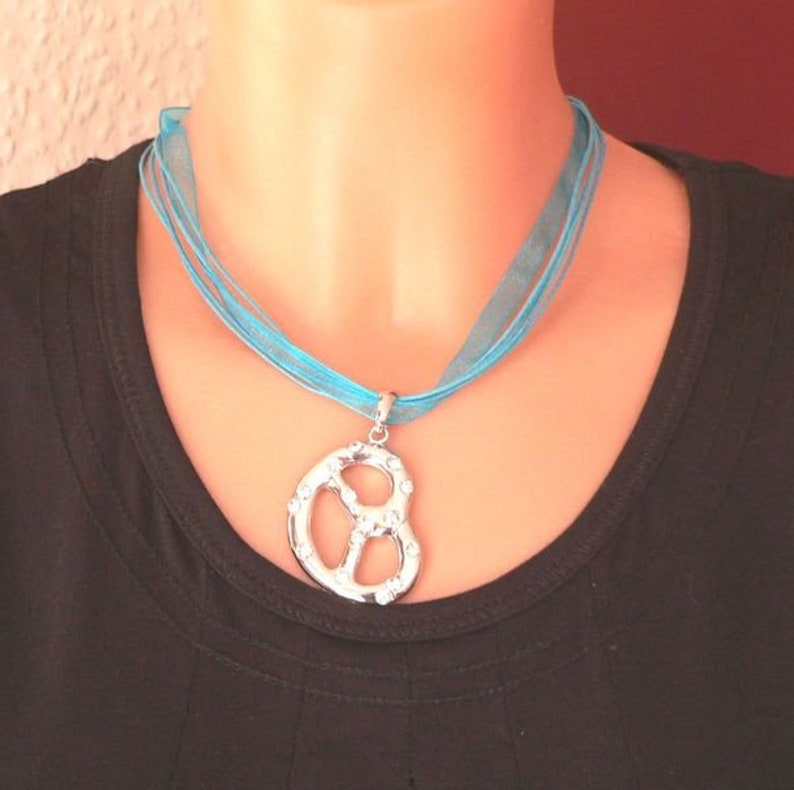 Peut inclure: Un collier en ruban turquoise avec un pendentif en forme de bretzel argent&eacute; incrust&eacute; de strass transparents.