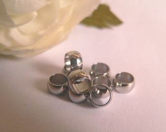 May include: A collection of shiny, silver-colored metal ring beads. The beads are round with a hole through the center. A white flower and green leaf are in the background. These beads are suitable for jewelry making and crafting.