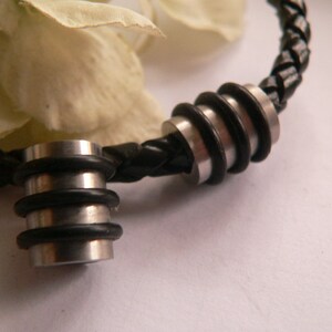 Op de afbeelding: Een close-up van een zwarte leren armband met twee zilveren metalen kralen. De kralen zijn rond en hebben een gegroefde textuur. De armband ligt op een wit oppervlak.