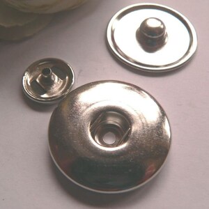 Op de afbeelding: Drie zilveren metalen drukknopen, een met een verhoogd midden, een met een verzonken midden en een met een plat midden.