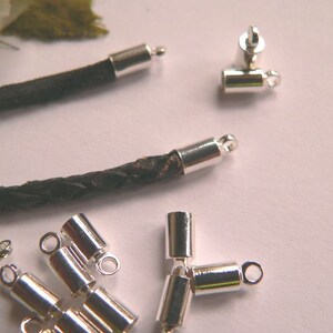 Può includere: Cappucci terminali per cordino in metallo argentato con un anello per il fissaggio a un cordino o una catena. I cappucci terminali per cordino sono mostrati su un cordino in pelle nera.