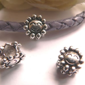 May include: Three silver-toned metal beads with a smiley face design. The beads are in the shape of a sun with a raised smiley face in the center. The beads are strung on a purple braided cord.