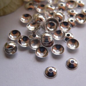 Può includere: Un gruppo di cappucci per perline in metallo color argento, sparsi su una superficie bianca. I cappucci sono rotondi con un piccolo foro al centro.