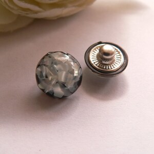 Könnte beinhalten: Zwei runde schwarze und weiße Knöpfe mit einem silbernen Metallkopf. Die Knöpfe haben einen Durchmesser von etwa 2,5 cm.