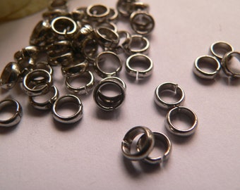May include: A pile of silver jump rings, a type of jewelry finding, scattered on a white surface. The rings are small and round, with a gap in the circle.