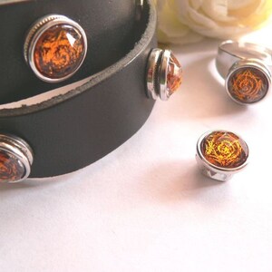Könnte beinhalten: Ein Lederarmband mit vier runden Druckknöpfen in Silber und Bernstein. Das Armband ist zweimal um sich selbst gewickelt. Die Druckknöpfe haben ein detailliertes Design.