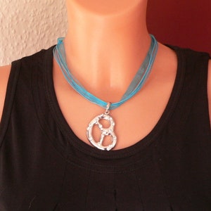 Peut inclure: Un collier ras de cou en ruban bleu turquoise avec un pendentif en forme de bretzel argent&eacute;. Le collier est port&eacute; par un mannequin avec un d&eacute;bardeur noir.