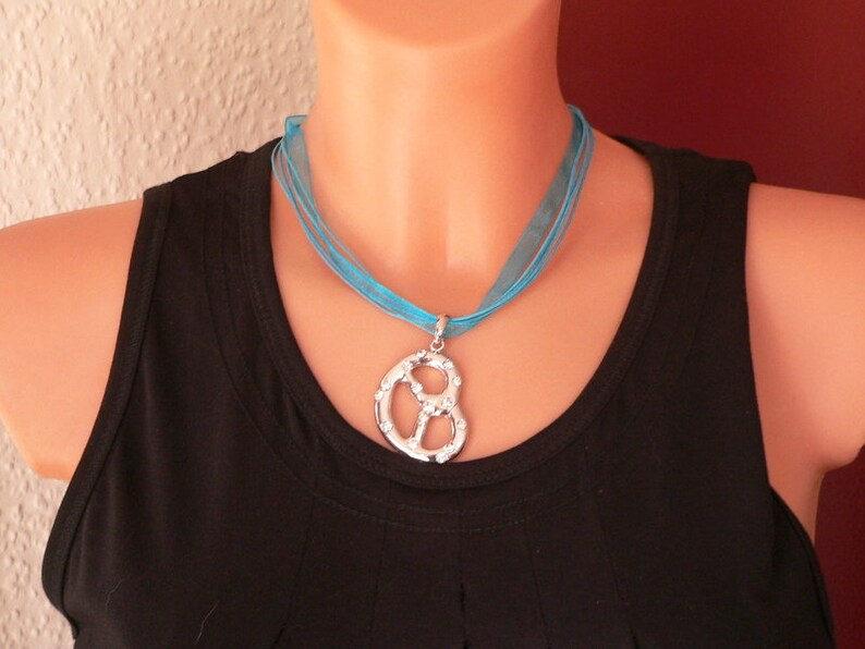Peut inclure: Un collier ras de cou en ruban bleu turquoise avec un pendentif en forme de bretzel argent&eacute;. Le collier est port&eacute; par un mannequin avec un d&eacute;bardeur noir.