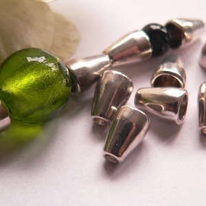 Può includere: Primo piano di tappi terminali in metallo argentato per la creazione di gioielli. I tappi terminali sono a forma di cono e hanno una finitura lucida. Nell'immagine è presente anche una perla di vetro verde.