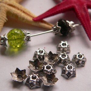 Peut inclure: Des embouts de perles en métal argenté avec un motif en étoile. Les embouts sont disposés sur une surface blanche avec une perle de verre verte et une perle noire.