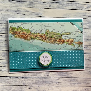 Könnte beinhalten: Handgefertigte Grußkarte mit einer Karte einer Insel. Die Karte hat einen türkisfarbenen Tupfenrand und eine kreisförmige Verzierung mit den Worten "Gute Reise". Die Karte liegt auf einer Holzoberfläche.