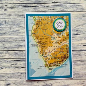 Könnte beinhalten: Eine weiße Karte mit blauem Rand zeigt eine Karte des südlichen Afrikas mit dem Text "Gute Reise" in einem blauen Kreis.
