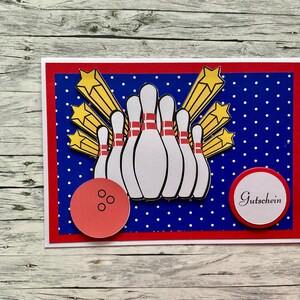Könnte beinhalten: Eine rechteckige Grußkarte mit blauem Hintergrund und weißen Punkten, einer Gruppe weißer Bowling-Pins mit roten Streifen, gelben Sternen, einer roten Bowlingkugel mit drei schwarzen Löchern und einem roten Kreis mit dem Text Gutschein.