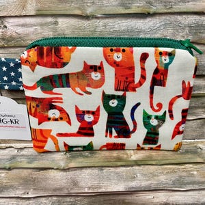 Könnte beinhalten: Ein kleines Stoffetui mit Reißverschluss und einem bunten Muster aus stilisierten Katzen in Orange, Rot, Grün und Blau auf cremefarbenem Grund. Das Etui hat einen dunkelgrünen Reißverschluss und eine blaue Lasche mit weißen Sternen.