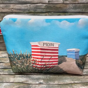 Könnte beinhalten: Eine Reißverschlusstasche mit einer Küstenszene, die einen rot-weiß gestreiften Strandkorb mit der Aufschrift MOIN, einen blau-weiß gestreiften Strandkorb, Sanddünen und einen blauen Himmel zeigt. Seitlich befindet sich eine rot-weiße Lasche mit Punkten.