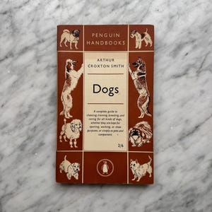 May include: A vintage book titled "Dogs" by Arthur Croxton Smith, part of the Penguin Handbooks series. The cover is a reddish-brown with illustrations of various dog breeds and the Penguin logo. The book provides a guide to choosing, training, and caring for dogs.