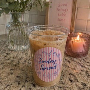 Puede incluir: Un vaso de plástico transparente lleno de café helado. El vaso tiene una pegatina circular con las palabras "Sunday Spread" e ilustraciones de alimentos. Una vela encendida y una impresión enmarcada con las palabras "good things take time" están en el fondo.