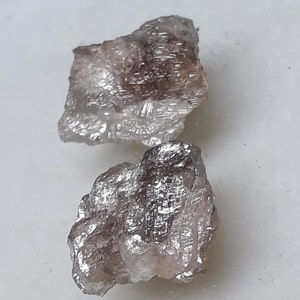 Op de afbeelding: Twee ruwe, onbewerkte, grijze diamanten. De diamanten hebben een zilveren glans en zijn onregelmatig gevormd.
