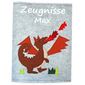 Può includere: Una cartella in feltro grigio con un design di drago marrone e rosso. La cartella ha il testo "Zeugnisse Max" sul davanti.