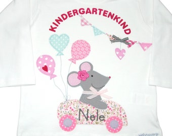 May include: White long-sleeved top featuring a mouse design. The mouse is in a floral car and holding balloons. The shirt has the text "KINDERGARTENKIND" in pink, and the name "Nele" is on the car.