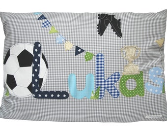 May include: A grey and white checked pillow with the name "Lukas" spelled out in colourful fabric letters. The pillow also features a football, a trophy, and a pair of football boots.