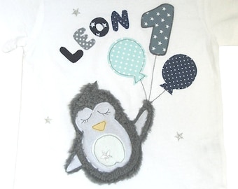 May include: White short-sleeved t-shirt with a grey penguin holding three balloons. The name "LEON" and the number "1" are above the penguin. The balloons are light blue, dark blue, and light blue with white polka dots. Small silver stars are scattered around the design.