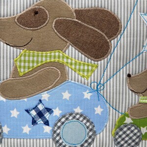 May include: Two brown felt dog appliques on a blue and white striped fabric. One dog is driving a blue car with white stars and a chequered wheel. The other dog is holding a green balloon with a white star.