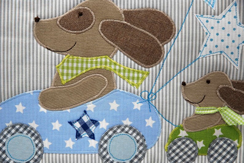 May include: Two brown felt dog appliques on a blue and white striped fabric. One dog is driving a blue car with white stars and a chequered wheel. The other dog is holding a green balloon with a white star.