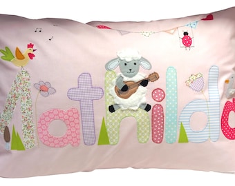 Op de afbeelding: Roze kussen met de naam "Matilda" in opgenaaide stoffen letters. Een schaap dat gitaar speelt staat centraal, met een haan, bloemen en andere decoratieve elementen.