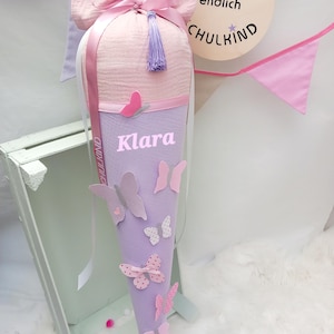 Könnte beinhalten: Ein lila-rosa Schultüte mit dem Namen "Klara" darauf. Die Tüte ist mit Schmetterlingen und einem rosa Quast verziert.