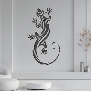 Op de afbeelding: Een donkergrijze muursticker van een hagedis in een tribaal ontwerp. De hagedis is afgebeeld met ingewikkelde patronen en een gekrulde staart. De sticker is aangebracht op een witte muur, wat een opvallend contrast creëert. Dit decoratieve stuk voegt een unieke touch toe aan elke kamer.