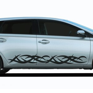 Op de afbeelding: Een zilveren auto met een zwarte tribal design sticker aan de zijkant. De sticker is een gestileerd patroon van gebogen lijnen en punten.