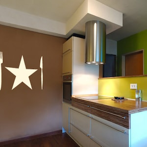 Può includere: Una stella bianca, una forchetta e un coltello sono su una parete marrone in una cucina. La cucina ha una cappa aspirante in acciaio inox, armadi in legno e un paraschizzi in vetro.