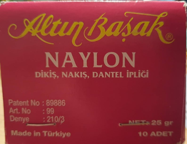 May include: A red box with gold lettering that reads "Altın Basak" and "NAYLON DİKİŞ, NAKIŞ, DANTEL İPLİĞİ". The box also includes the text "Patent No: 89886", "Art. No: 99", "Denye: 210/3", "NET: 25 gr", "Made in T&uuml;rkiye", and "10 ADET".