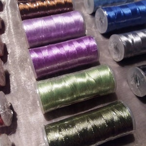 May include: Assortment of spools of thread in various colours, including brown, purple, green, and blue. The spools are wrapped in clear plastic and are arranged in a row.