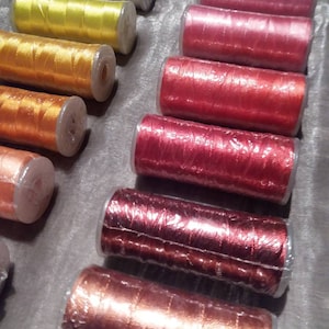 May include: Assortment of spools of thread in various colours, including silver, yellow, orange, red, and brown. The spools are wrapped in clear plastic.
