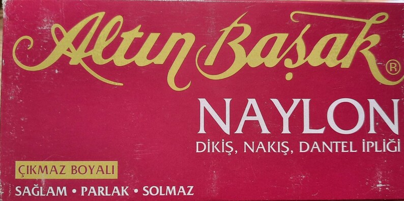 May include: A red box with gold lettering that reads "Altın Başak" and "NAYLON" with the text "DİKİŞ, NAKİŞ, DANTEL İPLİĞİ" below. The box also has the text "&Ccedil;IKMAZ BOYALI" and "SAĞLAM &bull; PARLAK &bull; SOLMAZ" on it.