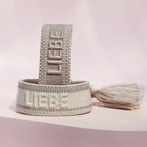 Könnte beinhalten: Zwei gewebte Armbänder in Beige mit dem in Weiß gestickten Wort "LIEBE". Das obere Armband ist ein dünneres Band und das untere Armband ist ein dickeres Band mit einem weißen Quasten.