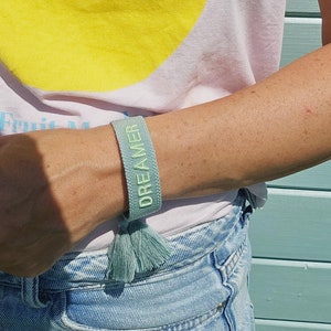 Könnte beinhalten: Ein hellblaues Armband mit einem weißen Quaste und dem Wort "DREAMER" in Grün gestickt.