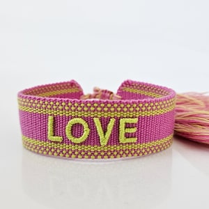 May include: A pink and yellow woven friendship bracelet with the word "LOVE" embroidered in yellow thread. The bracelet has a pink and yellow tassel.